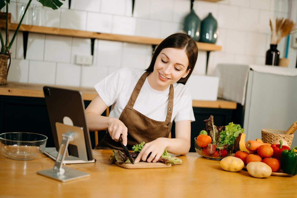 Jess Thompson happily chops fresh lettuce on a wooden board while looking at a recipe on a tablet in her bright, welcoming kitchen.
