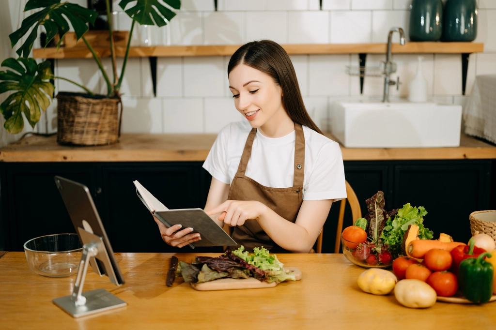 Jessica Thompson, a home cook in a brown apron, smiles as she reads a recipe book at her wooden kitchen table filled with fresh vegetables.