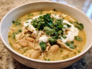 A close-up of a white bowl filled with creamy white chicken chili, topped with shredded chicken, white beans, sour cream, and fresh cilantro.