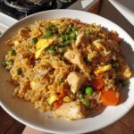 A close-up, brightly lit serving of amazing chicken fried rice with chunks of chicken, scrambled egg, peas, and carrots.