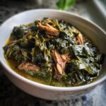 Close-up of a white bowl filled with tender, dark green collard greens simmered in broth with shredded smoked meat.