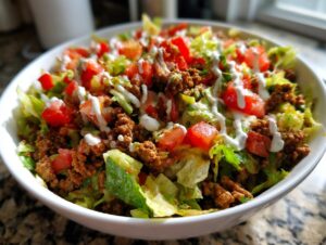A close-up view of a vibrant taco salad in a white bowl, featuring seasoned ground beef, crisp lettuce, diced tomatoes, and a drizzle of white dressing.