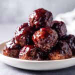 Close-up of a small white bowl piled high with glossy, saucy grape jelly meatballs.
