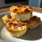 A stack of four golden brown, fluffy egg bites topped with crispy bacon bits, served on a white plate.