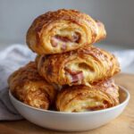 A stack of golden brown baked ham and cheese croissants cut open to show melted cheese and ham filling.