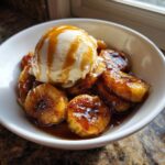A white bowl filled with caramelized bananas foster, topped with a scoop of vanilla ice cream drizzled with sauce.
