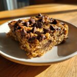 A close-up of a single, chewy oatmeal bars square topped generously with melted chocolate chips on a light plate.