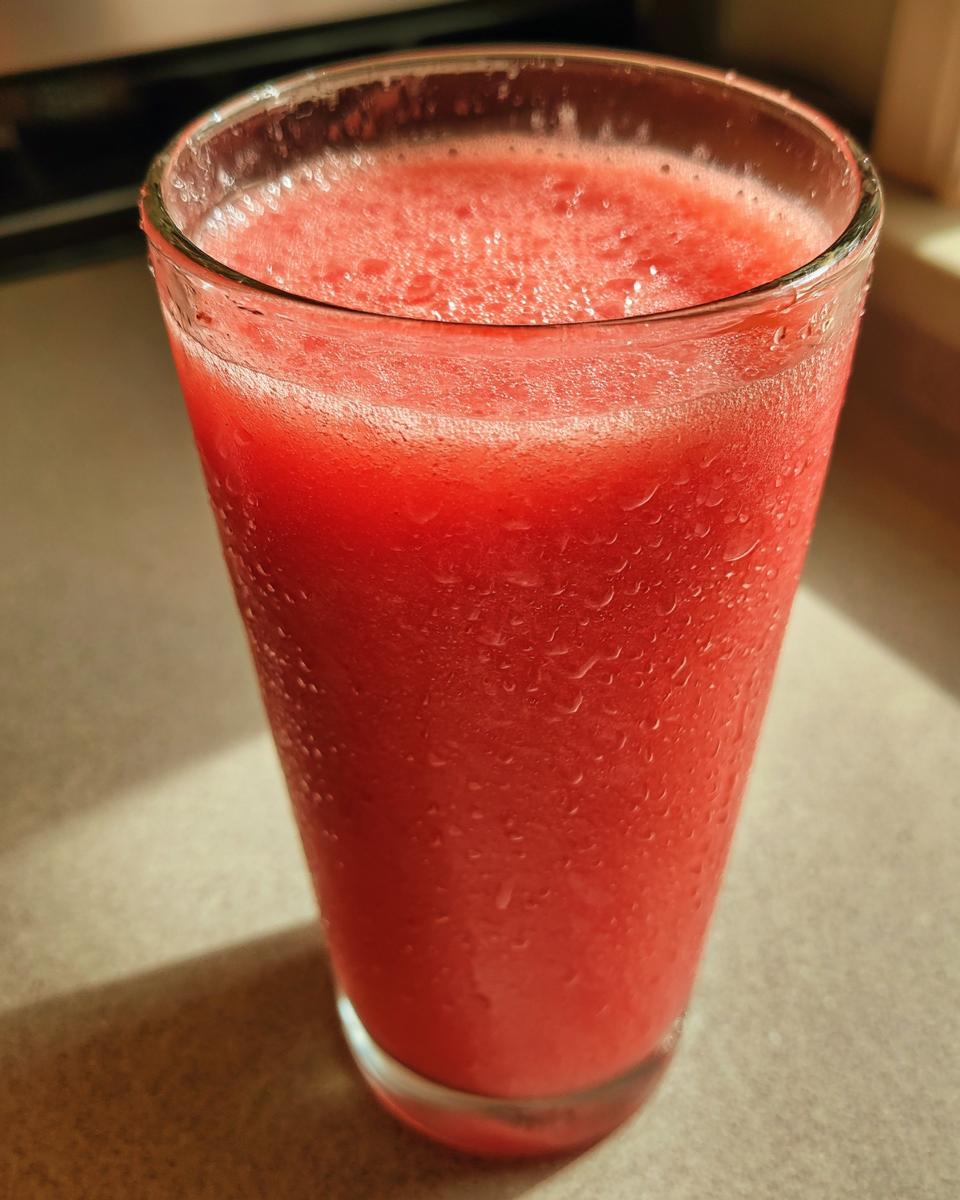 A tall, cold glass filled with a bright red, frothy watermelon smoothie, showing condensation droplets on the outside.