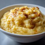 A close-up of creamy, golden cheese grits topped with a browned, bubbly cheese crust, served in a white bowl.