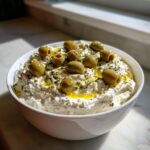 A white bowl filled with creamy dirty martini dip, garnished with whole green olives and olive oil.