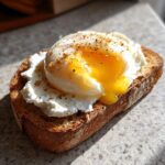 Close-up of creamy egg toast featuring a poached egg with a runny yolk over ricotta cheese on toasted bread, seasoned with pepper.