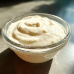 Close-up of thick, creamy homemade garlic sauce swirled in a small clear glass bowl.
