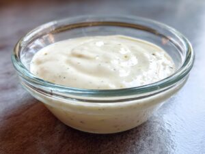 Close-up of a small glass bowl filled with thick, creamy homemade garlic sauce speckled with herbs.