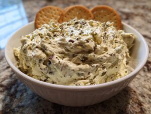 A bowl of creamy olive dip topped with herbs and served with three round crackers standing upright.