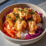 Close-up of a bowl filled with rice, crispy pieces of salmon, shredded carrots, purple cabbage, and drizzled with bang bang sauce.