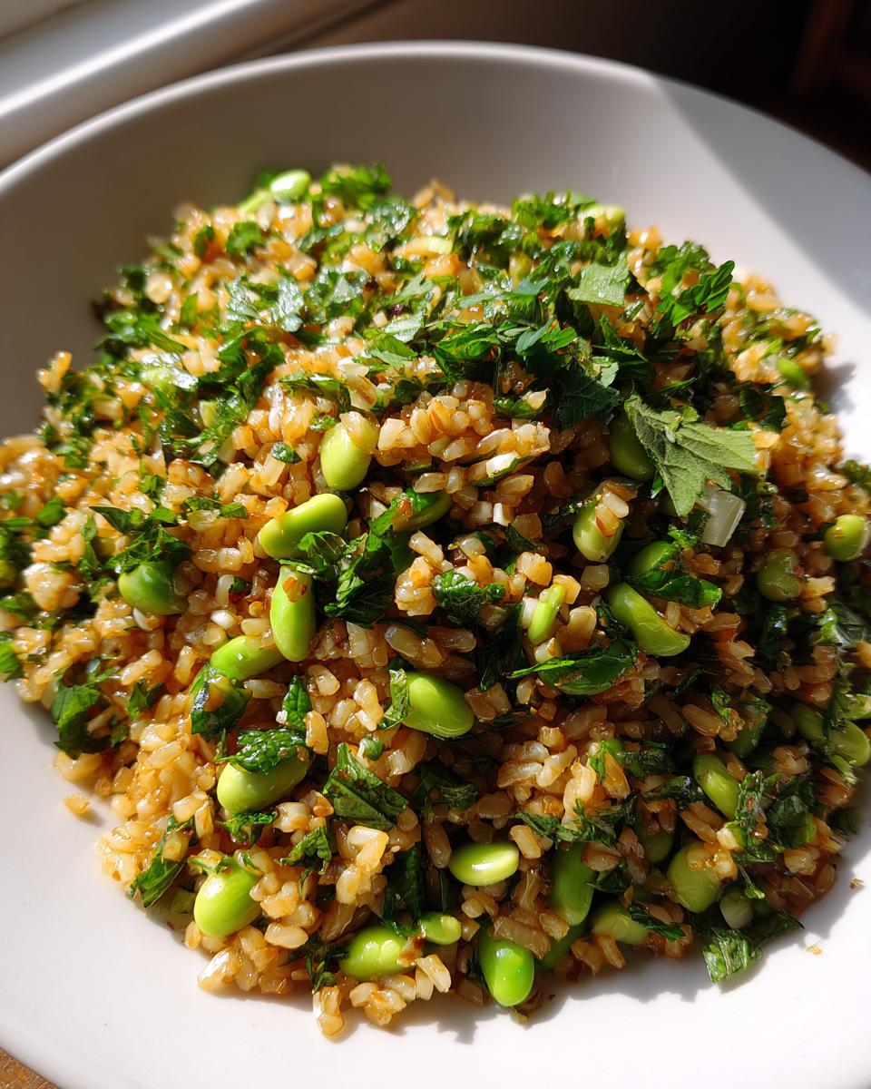 A close-up, sunlit bowl of crispy rice salad mixed with bright green edamame beans and chopped fresh herbs.