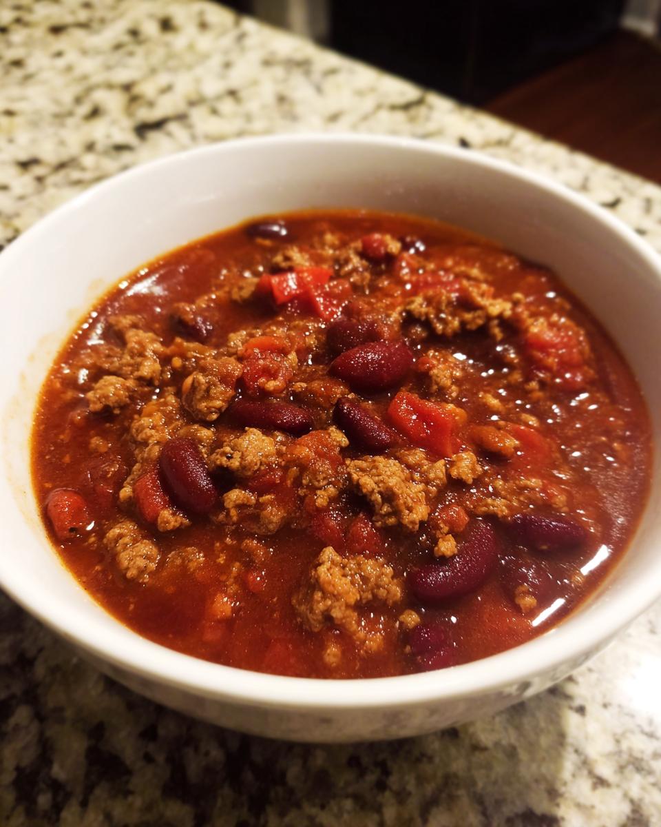 A close-up of a white bowl filled with rich, meaty crockpot chili featuring kidney beans and ground beef.