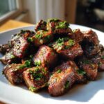 A pile of tender, browned crockpot steak bites seasoned with fresh herbs and garlic on a white plate.