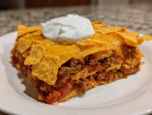 A close-up of a layered slice of Dorito Casserole topped with melted cheese, crunchy chips, and sour cream.