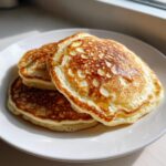 Close-up of three golden brown fluffy pancakes stacked on a white plate with a drizzle of syrup.