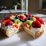 A cross-section view of a fruit pizza sugar cookie base topped with cream and fresh strawberries, kiwi, and blueberries.