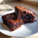 Two rich, fudgy pieces of sweet potato brownies stacked on a white plate in bright sunlight.