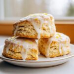 Three freshly baked lemon scones stacked on a plate, generously topped with dripping white lemon glaze and bright yellow zest.