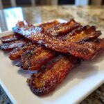 Close-up of several glistening, candied strips of Millionaire bacon piled on a white rectangular plate.
