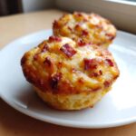 Close-up of two freshly baked pizza muffins topped with melted cheese and crispy bacon bits on a white plate.