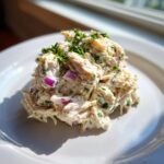 A mound of creamy greek yogurt chicken salad mixed with red onion and topped with fresh parsley on a white plate.