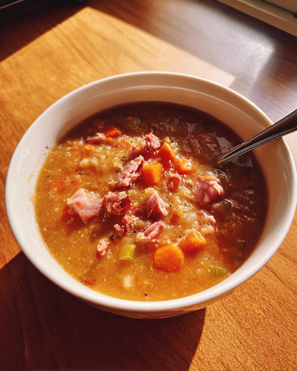 A close-up of a white bowl filled with thick, hearty Ham and Bean Soup, topped with chunks of ham and visible carrots.