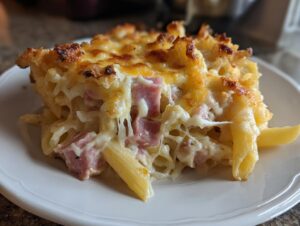 A close-up slice of baked ham and cheese casserole featuring penne pasta, diced ham, and a bubbly, browned cheese topping.