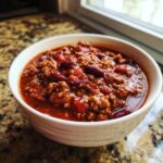A close-up view of a white bowl filled with rich, thick crockpot chili featuring ground beef and red kidney beans.