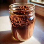 Close-up of rich brown hot cocoa mix stored in a clear glass mason jar, highlighted by sunlight.