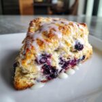 A close-up of a freshly baked, triangular blueberry scone topped with a sweet white glaze.