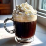 A clear glass mug filled with dark Irish Coffee topped with a generous layer of whipped cream.