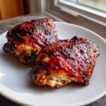 Two pieces of juicy, glazed bbq chicken thighs with a dark, caramelized sauce resting on a white plate.