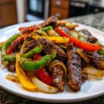 Close-up of seasoned steak fajitas mixed with vibrant red, green, and yellow bell peppers and onions on a white plate.