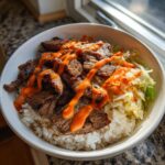 A white bowl filled with white rice, topped with grilled Korean BBQ steak and shredded cabbage, drizzled generously with spicy orange sauce.