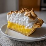 A tall slice of lemon meringue pie showing thick yellow curd and fluffy, toasted meringue peaks.
