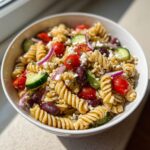 A close-up overhead shot of a vibrant mediterranean pasta salad featuring rotini pasta, cherry tomatoes, cucumbers, feta cheese, and olives.