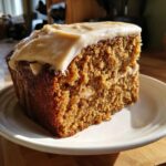 Close-up of a thick slice of moist applesauce cake topped with creamy frosting, sitting on a white plate.