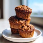 A stack of four dark, moist bran muffins with crumbly tops sitting on a small white plate.