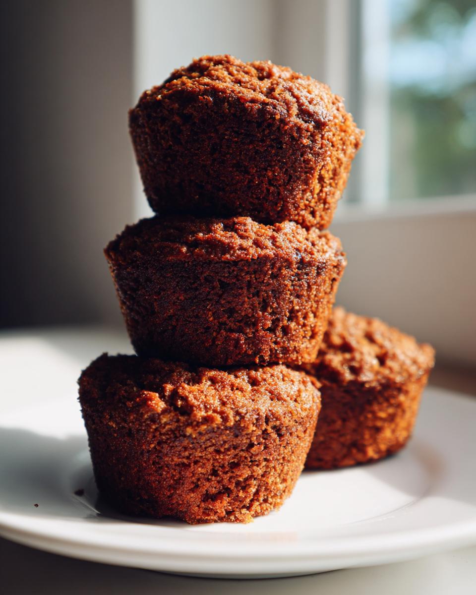 Close-up of three dark, moist bran muffins stacked on a white plate, highlighted by natural light.