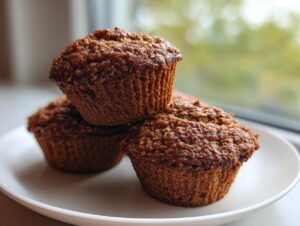 Three dark, moist bran muffins stacked on a white plate near a window.