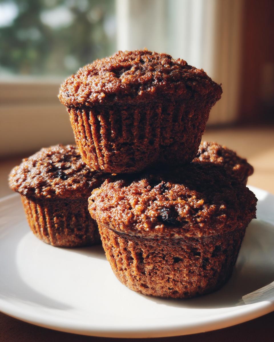 A stack of four moist, dark brown bran muffins sitting on a white plate near a sunlit window.