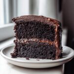A close-up of a moist, dark slice of chocolate cake with thick chocolate frosting, served on a white plate.