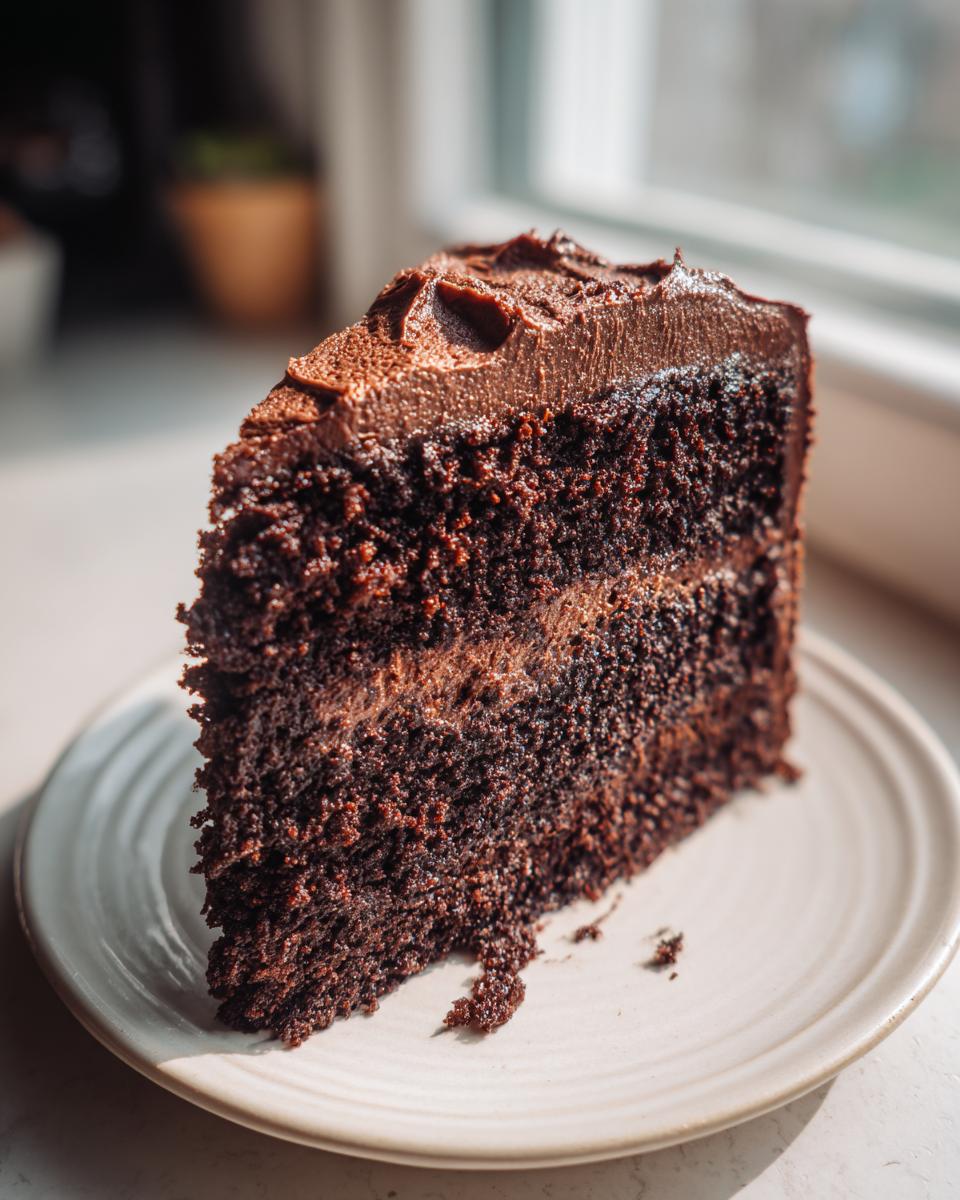 Close-up of a moist, dark slice of chocolate cake with thick chocolate frosting layers, sitting on a light plate.