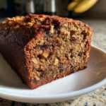 A close-up, appetizing shot of a moist slice of date nut bread filled with walnuts and dates, sitting on a white plate.