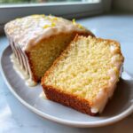 A slice cut from a loaf of moist lemon pound cake topped with a thick glaze and lemon zest.